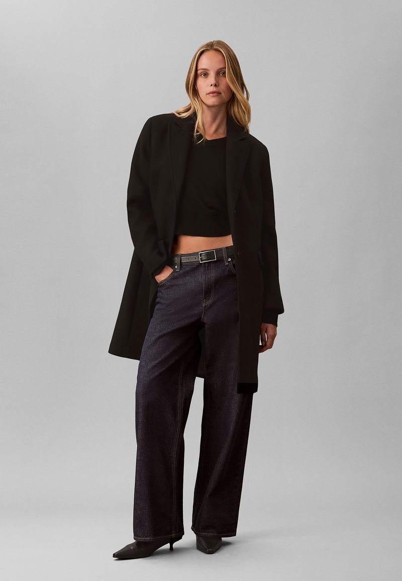 Femme aux longs cheveux blonds portant un long manteau noir, un crop top noir, un jean large foncé, une ceinture noire et des talons pointus noirs, debout devant un fond gris.