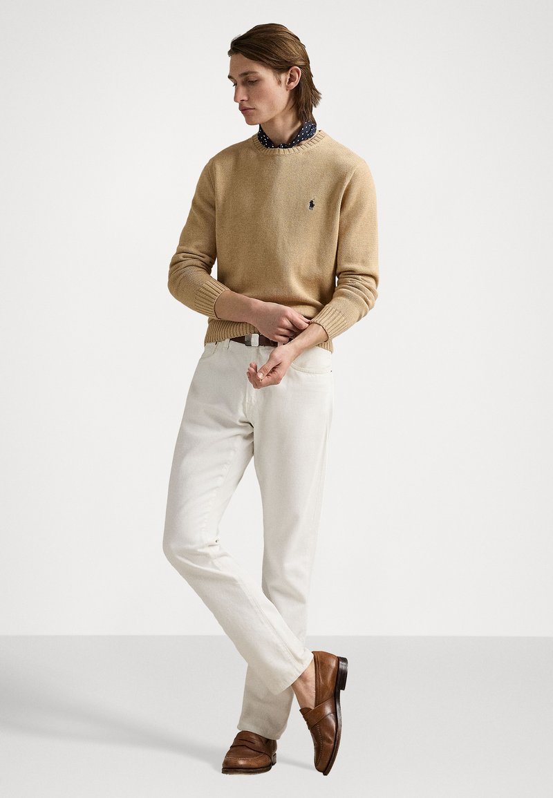 Beige Strickpullover mit gerippten Bündchen über einem navyblauen Polka-Dot-Hemd, kombiniert mit weißen Hosen und braunen Slippern. Minimalistisches Design, lockere Passform.
