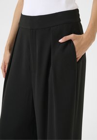 Pantalon noir à jambes larges avec une texture lisse, doté d'une taille haute et de poches latérales, complété par de subtils plis à l'avant.