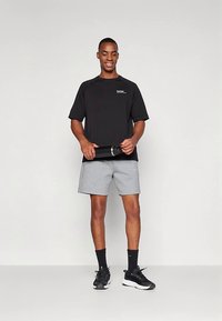 Schwarzes kurzärmliges T-Shirt mit Logo, graue Shorts und schwarze Turnschuhe. Das Modell hält eine schwarze Wasserflasche und steht vor einem neutralen Hintergrund.