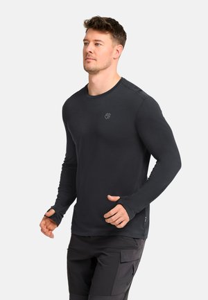Man wearing a fitted black long-sleeve shirt with thumb holes and black cargo pants, standing against a plain light background.