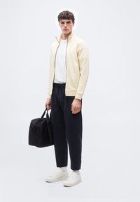 Νέος άνδρας με ανοιχτό μπεζ τζάκετ με φερμουάρ, λευκό μπλουζάκι, μαύρο παντελόνι, λευκά αθλητικά παπούτσια, κρατώντας μαύρο σακ βουαγιάζ, στέκεται σε απλό φόντο.