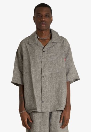 Uomo che indossa una camicia grigia a maniche corte, ampia, con bottoni e pantaloni abbinati, in piedi davanti a uno sfondo semplice.