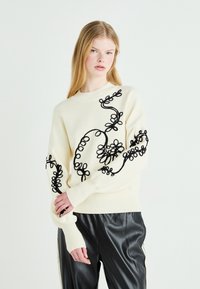 Crewneck genser i kremfarge med svart blomsterbroderi. Har lange ermer og ribbestrikkede mansjetter. Kombinert med svarte blanke skinnbukser.