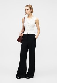 Frau mit kurzen blonden Haaren, die ein ärmelloses weißes Oberteil mit Rüschen, schwarze weite Hosen, schwarze Schuhe trägt und eine braune Schultertasche bei sich hat.