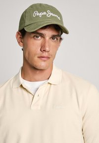 Olīvzaļš cepure ar baltu "Pepe Jeans" izšuvumu, kombinēta ar krēmkrāsas polo kreklu, kurā ir smalks logotips. Gluda auduma tekstūra, ikdienišķs dizains.