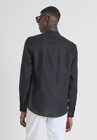 Camicia nera a maniche lunghe con colletto, tessuto testurizzato e polsini con bottoni, indossata con pantaloni bianchi, vista da dietro.