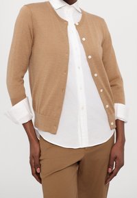 Beige pletený kardigan s kulatým výstřihem, zapínáním na knoflíky a tříčtvrtečními rukávy, nošený přes bílou košili s límečkem.