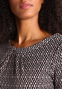 Femme aux cheveux bouclés et foncés portant un haut à motifs noirs et blancs avec un col rond, visible du cou jusqu'à la partie supérieure de la poitrine.