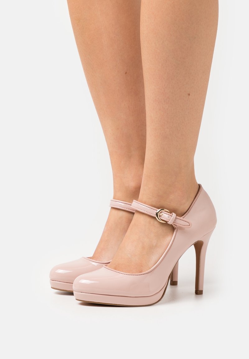 Escarpins Mary Jane brillants rose clair avec brides à la cheville et boucles dorées, portés pieds nus sur fond blanc.