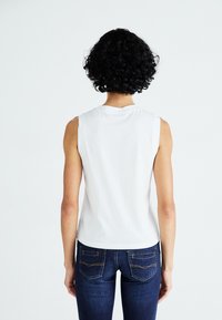 Hvit ermeløs topp med rund hals, sammen med mørkeblå jeans med gule sømmer og detaljer på baklommene.