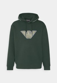 Grön huvtröja med en stor, broderad logotyp i beige och svart i mitten. Har en dragsko vid huvan och ribbade muddar.