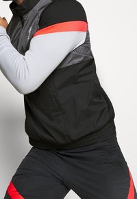 Chaqueta negra y gris con acentos rojos, con un alto cuello y una cintura elástica; combinada con pantalones oscuros a juego.