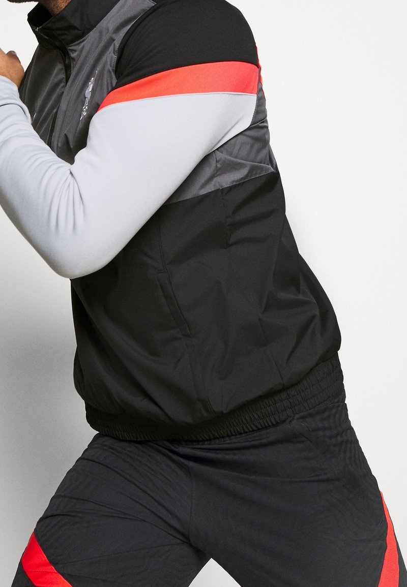 Chaqueta negra y gris con acentos rojos, con un alto cuello y una cintura elástica; combinada con pantalones oscuros a juego.