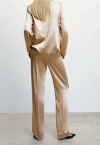 Krémový saténový oblek s upraveným sakem a širokými kalhotami. Vyznačuje se hladkým povrchem a čistými liniemi. Dlouhé vlasy spadají přes záda saka.