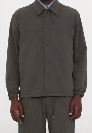Mann trägt dunkelgraue, geknöpfte Jacke mit elastischen Bündchen über hellblauem Hemd mit Kragen und passenden dunkelgrauen Hosen.