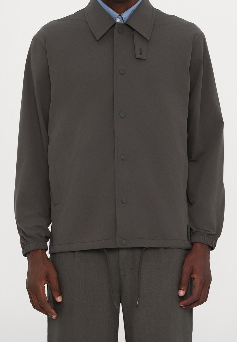 Homme portant une veste gris foncé boutonnée avec des poignets élastiques par-dessus une chemise bleu clair à col et un pantalon gris foncé assorti.