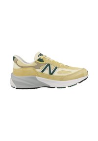 Zapatilla deportiva amarilla y beige con materiales de malla y ante, con acentos verdes y un logo blanco, mediasuela gris curvada y suela negra.