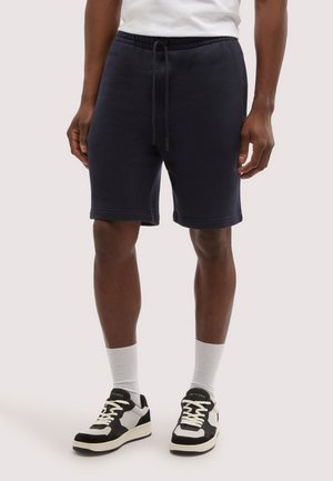 Mężczyzna ubrany w czarne szorty ze ściągaczem, białe skarpety do kostek i czarno-białe trampki, stojący na tle jednolitego jasnego tła.