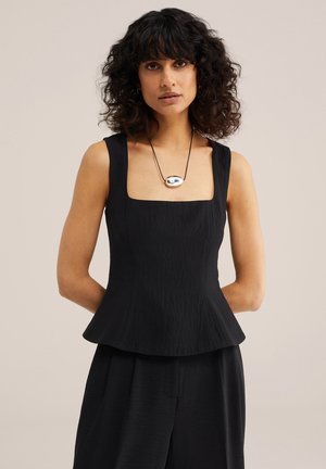 Kobieta o kręconych ciemnych włosach, ubrana w czarną, bezrękawnikową bluzkę peplum i czarne spodnie, ozdobiona srebrnym naszyjnikiem z zawieszką.