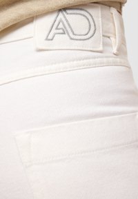 Białe dżinsy z fakturowanym wykończeniem. Posiadają naszywkę z napisem "AD" w szarym przeszyciu z tyłu. Zawierają tylną kieszeń i szlufki na pasek.
