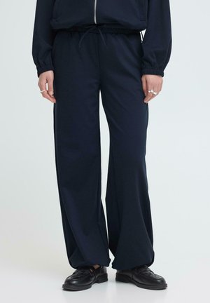 Osoba stojaca pred svetlým jednoduchým pozadím, oblečená v voľných čiernych nohaviciach so sťahovacou šnúrkou v páse a čiernych kožených topánkach.