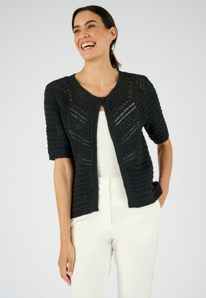 Mujer sonriente que lleva un cárdigan de punto negro de manga corta sobre una camiseta blanca y pantalones blancos, de pie con una mano en el bolsillo.