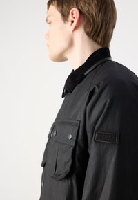 Black waterproof jacket featuring a corduroy collar, snap buttons, and a patch on the shoulder. Includes a front pocket.