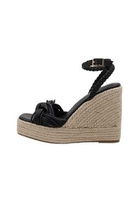 Schwarze Keil-Sandale mit einem geflochtenen Knöchelriemen, geknotetem Vorderdetail und einer strukturierten Jute-Plattform. Glattes schwarzes Obermaterial und natürlicher strukturierter Boden.