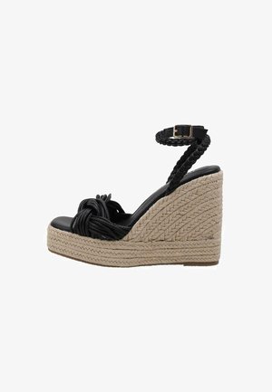 Sandale en wedge noire avec une sangle de cheville tressée, un détail noué à l'avant et une plateforme en jute texturée. Dessus lisse noir et base texturée naturelle.