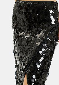 Falda negra de lentejuelas con una abertura alta, que presenta lentejuelas circulares que crean una superficie texturizada y reflejan la luz. Tela suave debajo.