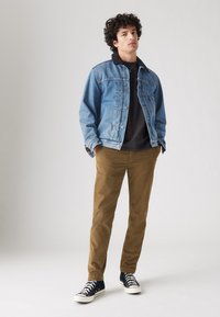 Chaqueta de mezclilla con cuello negro, camiseta negra, pantalones marrones y zapatillas negras. Los detalles notables incluyen cierre de botones y ajuste casual.