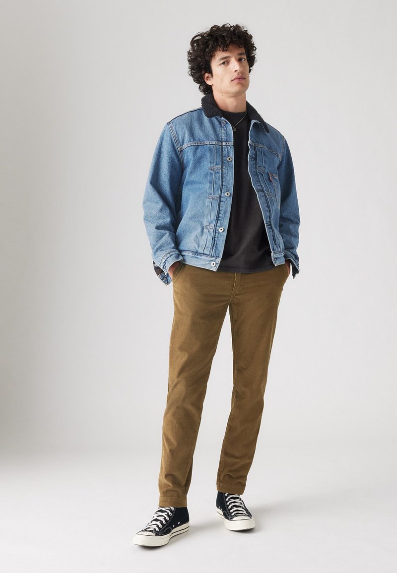 Veste en denim avec col noir, t-shirt noir, pantalon marron et baskets noires. Détails notables : fermeture à bouton et coupe décontractée.