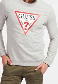 Szara bluza z tkaniny bawełnianej, z dużym czerwonym trójkątnym logo z napisem "GUESS U.S.A." i znakiem zapytania pośrodku.