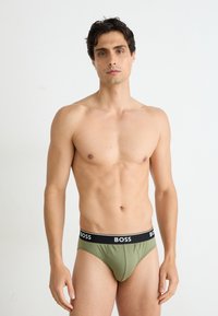 Shorts verts avec une ceinture logo BOSS noire, offrant un ajustement ajusté et une texture lisse, présentés sur un modèle sur un fond neutre.