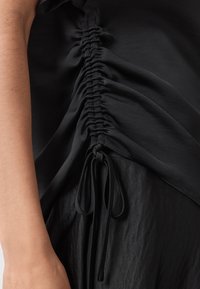 Top nero con gathered ai lati, con arricciature e lacci regolabili. Il tessuto ha una consistenza morbida e si drappeggia dolcemente.
