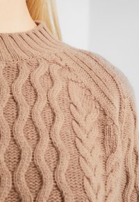 Beżowy sweter wykonany na drutach, z wypukłymi wzorami warkoczy oraz ściągaczem przy kołnierzu, uszyty z miękkiej mieszanki wełny o strukturalnej powierzchni.