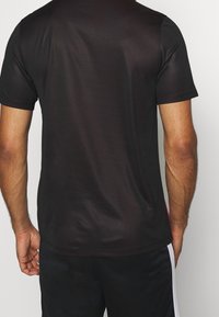 Hombre vestido con una camiseta negra de manga corta sencilla y pantalones cortos negros con rayas blancas en los costados, visto desde atrás contra un fondo liso.