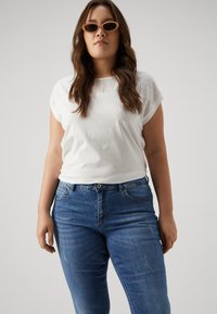White short-sleeve top with eyelet detail at the shoulders and light blue denim jeans. The model wears brown oval sunglasses.