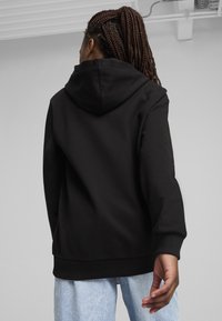 Felpa nera con cappuccio regolabile, polsini e orlo a costine, materiale in cotone morbido e vestibilità comoda, indossata con jeans azzurri chiari.