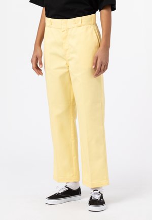Persona que lleva pantalones rectos de color amarillo claro, camiseta negra, calcetines blancos y zapatillas negras de corte bajo con cordones blancos.