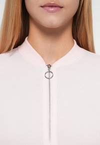 Veste rose clair à fermeture éclair avec un col rond, dotée d'une fermeture éclair argentée et d'un tire-zip en boucle. Tissu doux et extensible, texture lisse.