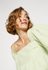 Top verde claro com ombros à mostra, mangas bufantes, cintura franzida e textura suave. Detalhe de costura mínima em uma cor sólida.