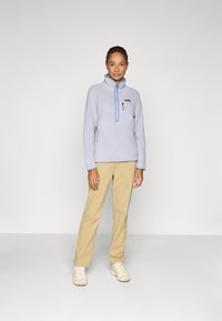 Lichtpaarse fleece pullover met een rits aan de hals, twee zijzakken en lichtblauwe accenten, gecombineerd met beige broek en lichte schoenen.