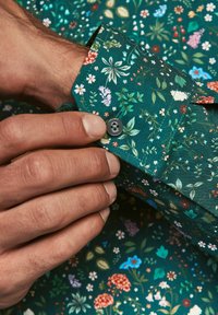 Mâneca cămășii cu imprimeu floral verde, dotată cu un nasture gri, având diverse flori colorate și frunze pe un material texturat. O mână ajustează mâneca.