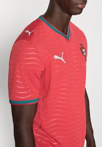 Κόκκινη ποδοσφαιρική φανέλα από υφασμάτινη υφή με V-λαιμόκοψη. Διαθέτει ανοιχτοχρωματικά κυματιστά μοτίβα, πράσινες και κόκκινες λεπτομέρειες στα μανίκια και λευκό λογότυπο στον ώμο.