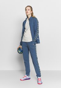 Niebieska sportowa kurtka z rękawami w wzór chevron, w zestawie z dopasowanymi spodniami. Lekki materiał, trzymający niebieską piłkę nożną. Wielokolorowe buty.