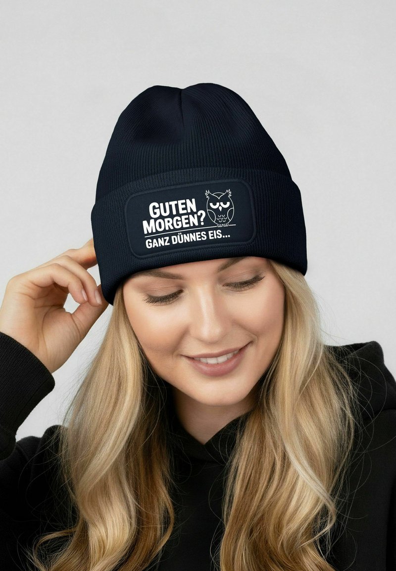 Black ribbed beanie with a rectangular patch displaying white text and an owl graphic. The fabric appears soft and flexible.
