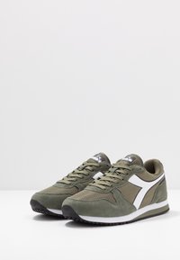 Zapatillas en verde olivo con materiales de malla y ante, acentos blancos, cordones redondos, cuello acolchado y suela de goma negra con buena tracción.