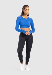 Donna in piedi con le mani congiunte, che indossa un crop top blu a maniche lunghe, leggings neri e sneakers bianche su uno sfondo grigio.